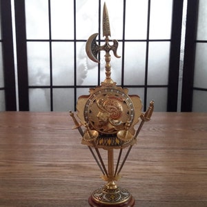 May include: Gold-toned metal decorative stand with a shield design and four swords. The shield features a knight's helmet and a gold and black design.