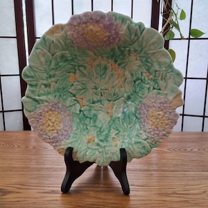 Peut inclure: Une assiette décorative en céramique avec un motif floral. L'assiette présente un motif en relief de feuilles et de fleurs dans des tons de vert, de jaune et de violet. Elle est présentée sur un présentoir en bois noir.