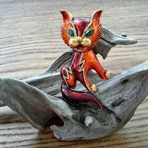 Peut inclure: Une broche en émail rouge et or en forme de chat avec des yeux verts et un motif d'éclair. La broche est posée sur un morceau de bois flotté.