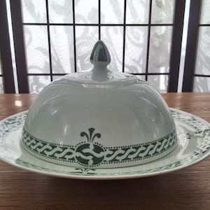Peut inclure: Un plat à beurre en céramique blanche avec un motif vert et blanc. Le plat a un couvercle bombé avec un bouton sur le dessus.