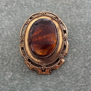 Peut inclure: Une broche de forme ovale avec un cabochon central translucide de couleur ambre orné d'un motif floral gravé. La pierre est sertie dans un cadre en métal doré vieilli décoratif présentant des détails complexes en filigrane et perlés.
