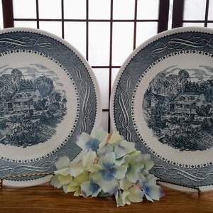 May include: Two blue and white ceramic dinner plates featuring a black and white illustration of a house in a wooded setting. The plates have a decorative blue border.