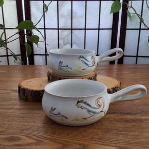 May include: Two white ceramic soup bowls with handles, each decorated with a whimsical design of a running rabbit and a rooster. The bowls are set on a wooden surface, showcasing a rustic, handmade aesthetic.