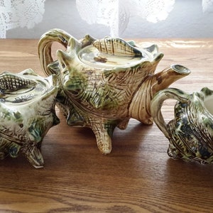 May include: A ceramic tea set with a teapot, creamer, and sugar bowl. The set is decorated with a green and brown seashell design.