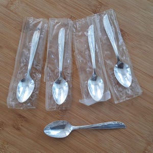 May include: Five silver teaspoons in individual plastic packaging, with one additional silver teaspoon lying on a wooden surface.