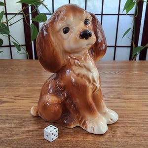 Peut inclure: Figurine en céramique représentant un chien assis, avec une fourrure marron et des accents crème. Le chien a de grandes oreilles et un nez noir. Un dé blanc à points noirs est devant le chien.