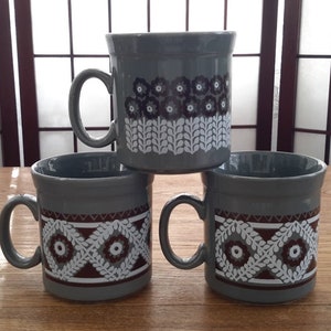 May include: Three grey ceramic mugs with brown and white geometric designs. The mugs have a rounded shape and a handle on the side.