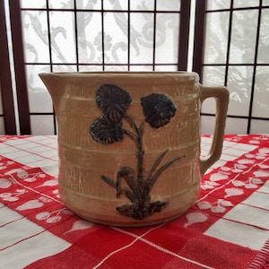 May include: A vintage brown stoneware pitcher with a blue and black floral design. The pitcher has a wide mouth and a handle.