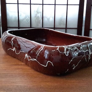 Peut inclure: Un pot en céramique brun avec un glaçage craquelé blanc. Le pot est de forme ovale et a une base large et peu profonde.