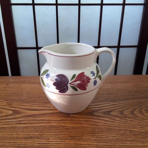 Peut inclure: Pichet en céramique blanche avec un motif floral dans les tons de rouge, violet, bleu et vert. Le pichet a un corps arrondi et une anse.