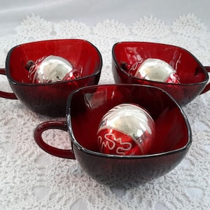 Peut inclure: Trois tasses en verre rouge avec des bords carrés et des poignées. Chaque tasse contient une boule de Noël argentée.