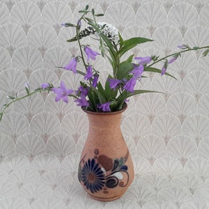 May include: A brown ceramic vase with a floral design, filled with purple bellflowers and green foliage. The vase has a dark blue and white floral pattern. The background is a white, textured fabric.