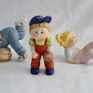 Peut inclure: Trois figurines en porcelaine d'enfants. Un enfant porte une casquette de baseball bleue avec les lettres "CP" dessus, une chemise rayée jaune et verte, une salopette rouge et un gant de baseball marron. Un autre enfant porte une chemise rayée bleu et blanc, une salopette bleue et fait un poirier. Le troisième enfant porte une robe rose et a des tresses blondes.