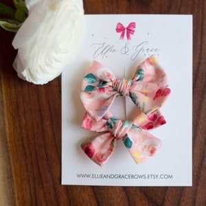 Könnte beinhalten: Zwei rosa florale Haarbänder mit grünen Blättern und weißen Blüten. Die Schleifen befinden sich auf einer weißen Karte mit dem Text "Ellie & Grace Bows" und einer Website-Adresse.