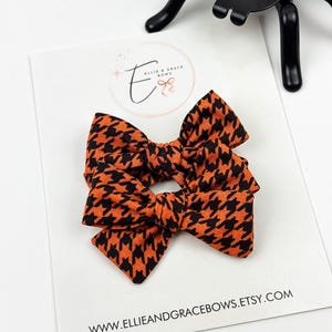 Op de afbeelding: Twee kleine, oranje en zwarte haarstrikken met een hondenpootpatroon. De strikken liggen op een witte kaart met de tekst "ELLIE & GRACE BOWS" en het websiteadres. Een zwarte spinnendecoratie staat op de achtergrond.