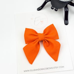 Può includere: Fiocco in tessuto arancione brillante su una carta bianca. Il fiocco è centrato sulla carta, con l'indirizzo del sito web www.ellieandgracebows.etsy.com stampato in basso. Un portacandele nero a forma di ragno sullo sfondo.