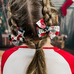 Peut inclure: Une jeune fille avec de longs cheveux bruns coiffés en tresses avec des nœuds en tissu rouge et vert sur le thème de Noël. Elle porte un t-shirt de baseball rouge et blanc.