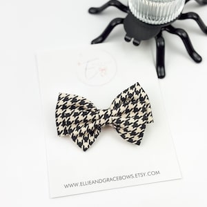 Può includere: Un fiocco nero e crema a motivo pied-de-poule su una carta bianca. Il fiocco è centrato sulla carta, con l'indirizzo del sito web www.ellieandgracebows.etsy.com stampato sotto. Un portacandele nero a forma di ragno è sullo sfondo.