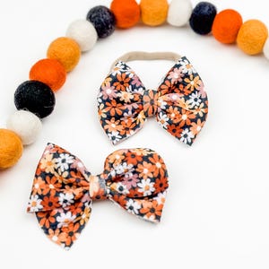 May include: Two floral print hair bows with a black background and orange, pink, and white flowers. The larger bow is on a nude elastic headband.