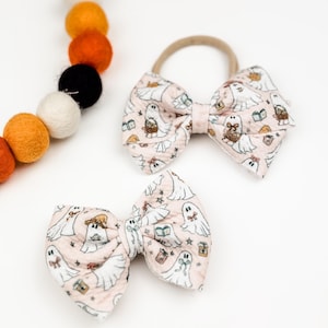 May include: Two pink and white hair bows with a ghost and Halloween themed print. The bows are tied with a nude elastic headband.