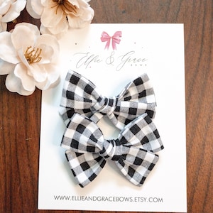 Peut inclure: Deux nœuds pour cheveux en vichy noir et blanc sur une carte blanche avec le texte "Ellie & Grace Bows". Les nœuds sont disposés sur une surface en bois avec des fleurs blanches en arrière-plan.