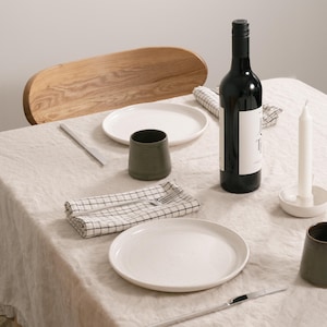 May include: A dining table setting featuring a beige linen tablecloth, two white ceramic plates, two dark green ceramic cups, two silver knives, a silver fork, a black wine bottle with a white label, and a white candle in a small white holder.