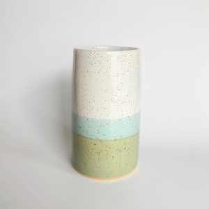 May include: A cylindrical ceramic vase featuring a speckled finish. The design is divided into three horizontal colour bands: a top section in off-white, a middle section in light turquoise, and a bottom section in sage green, all with dark speckles.