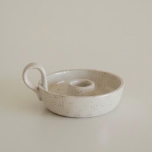 Peut inclure: Un petit bougeoir en céramique blanc cassé de forme ronde avec une texture mouchetée. L'objet présente un bassin bas et peu profond, une ouverture circulaire centrale pour une bougie et une anse incurvée fixée sur le côté du rebord.