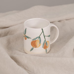 May include: White ceramic mug with a painted floral design of orange fruit and green leaves. The mug has a rounded body, a handle, and a slightly flared base. The design is hand-painted in a whimsical style.