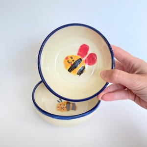 Peut inclure: Deux bols en céramique de couleur crème avec un bord bleu foncé. Chaque bol présente une illustration peinte à la main d'une abeille jaune et noire avec des ailes rouges au centre. Les bols ont une finition lisse et brillante.