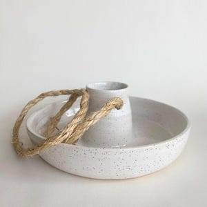 May include: A white ceramic serving bowl with a speckled finish, featuring a central cylindrical holder and a woven rope handle. The bowl has a wide, shallow design, ideal for serving snacks or displaying decorative items.