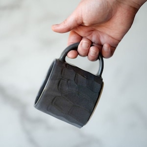 Peut inclure: Une tasse en céramique gris foncé avec une surface texturée et une anse arrondie. La tasse est tenue dans une main, mettant en valeur son design et son savoir-faire uniques. L'intérieur de la tasse a un bord de couleur claire.