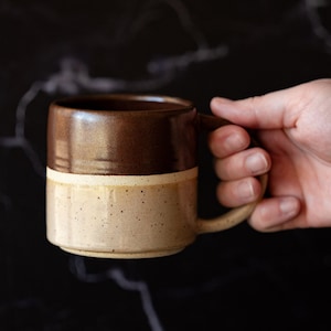 May include: A ceramic mug featuring a two-tone design with a glossy dark brown upper section and a speckled light beige lower section. The mug has a cylindrical body, a flat base, and a curved handle extending from the side of the vessel.