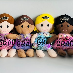 May include: Four plush dolls dressed in iridescent pink, purple, and blue dresses with gold stars and the text "Preschool Grad" on the front. Each doll has a brown, tan, yellow, or black head with a graduation cap on top. The caps have the year 2023 embroidered on them.