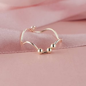May include: A gold-colored ring with a wavy design and small spherical accents. The ring's band has a flowing, organic shape, and the spheres are evenly spaced. The ring is displayed on a pink background, creating a soft and elegant aesthetic.