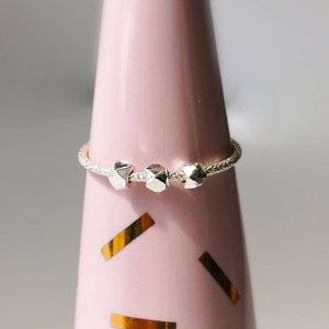 May include: A silver ring with three textured, geometric beads. The ring is on a pink display stand.
