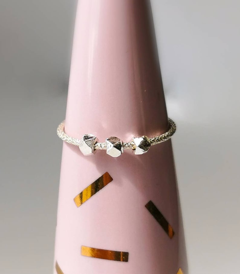 May include: A silver ring with three textured, geometric beads. The ring is on a pink display stand.
