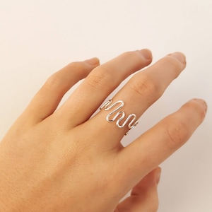 May include: A silver ring featuring an abstract, wavy design. The ring is crafted from a single, thin wire bent into a series of connected curves. The ring is worn on a finger, set against a plain, light-coloured background.