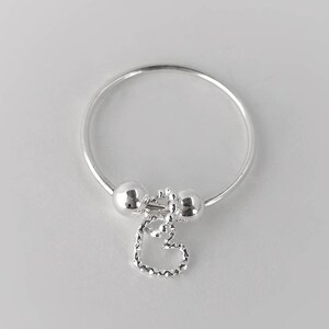 May include: A silver ring with two small silver balls and a dangling heart charm. The heart charm is made of small silver beads.