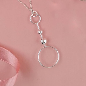 May include: A silver necklace with a long pendant featuring three round beads and two circles. The necklace is on a pink background.