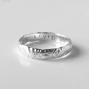 May include: A silver ring featuring an engraved floral pattern and the inscription "OUR LOVED". The ring has a detailed design with a slightly textured surface. The ring is a band style, suitable for everyday wear.