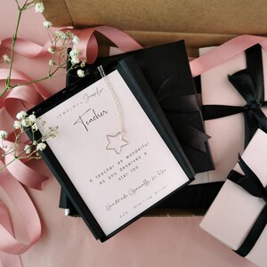 May include: A gift box with a pink and white ribbon, a black box containing a silver necklace with a star charm, and a card that reads "Teacher" and "A teacher as wonderful as you deserves a star too".