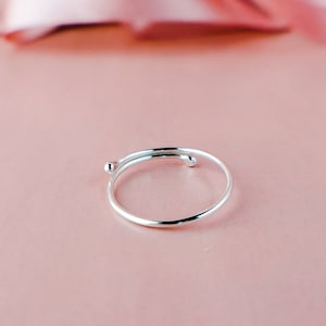 May include: A silver ring with a twisted design and a small ball on each end. The ring is on a pink surface.