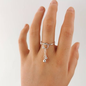 May include: A silver ring with a dangling chain and two small silver balls. The ring is on a person's finger.