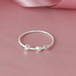 May include: A silver ring with three textured beads. The ring is on a pink background.