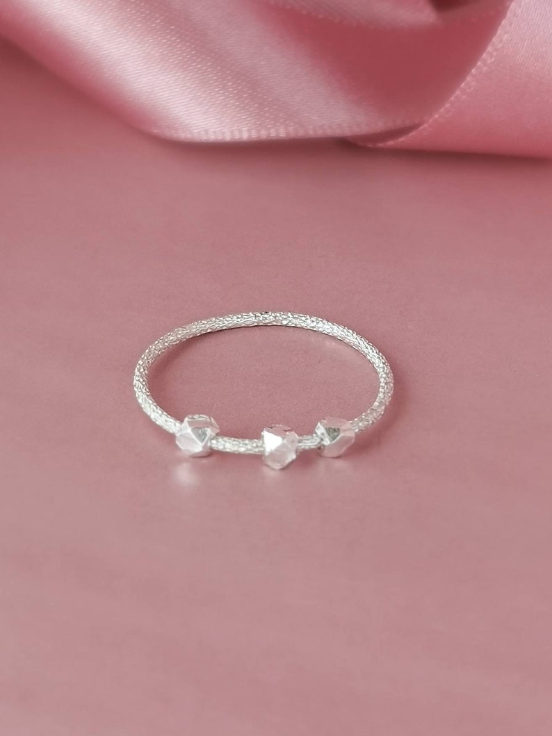 May include: A silver ring with three textured beads. The ring is on a pink background.
