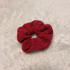 May include: A circular hair scrunchie crafted from deep red yarn with a visible crochet texture. The accessory features a gathered, ruffled design with a central opening, showcasing a consistent dark red colour throughout the entire fabric construction.