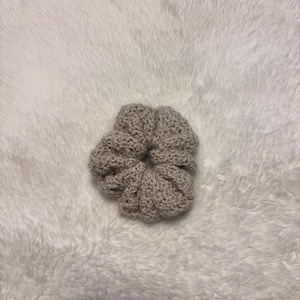May include: A light beige, circular hair scrunchie featuring a textured, crocheted yarn construction. The accessory has a gathered, ruffled appearance with a central opening, showcasing a consistent neutral colour and a soft, looped fibre finish.