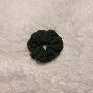 May include: A dark charcoal grey hair scrunchie featuring a textured, crocheted fabric construction. The circular accessory has a gathered, ruffled appearance with a central opening, showcasing a dense, uniform stitch pattern throughout the design.