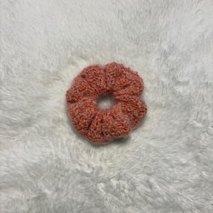 May include: A round, crocheted hair scrunchie featuring a textured, variegated yarn in shades of coral, peach, and white. The accessory has a gathered, ruffled appearance with a circular opening in the centre, showcasing a dense, looped stitch pattern throughout the entire piece.